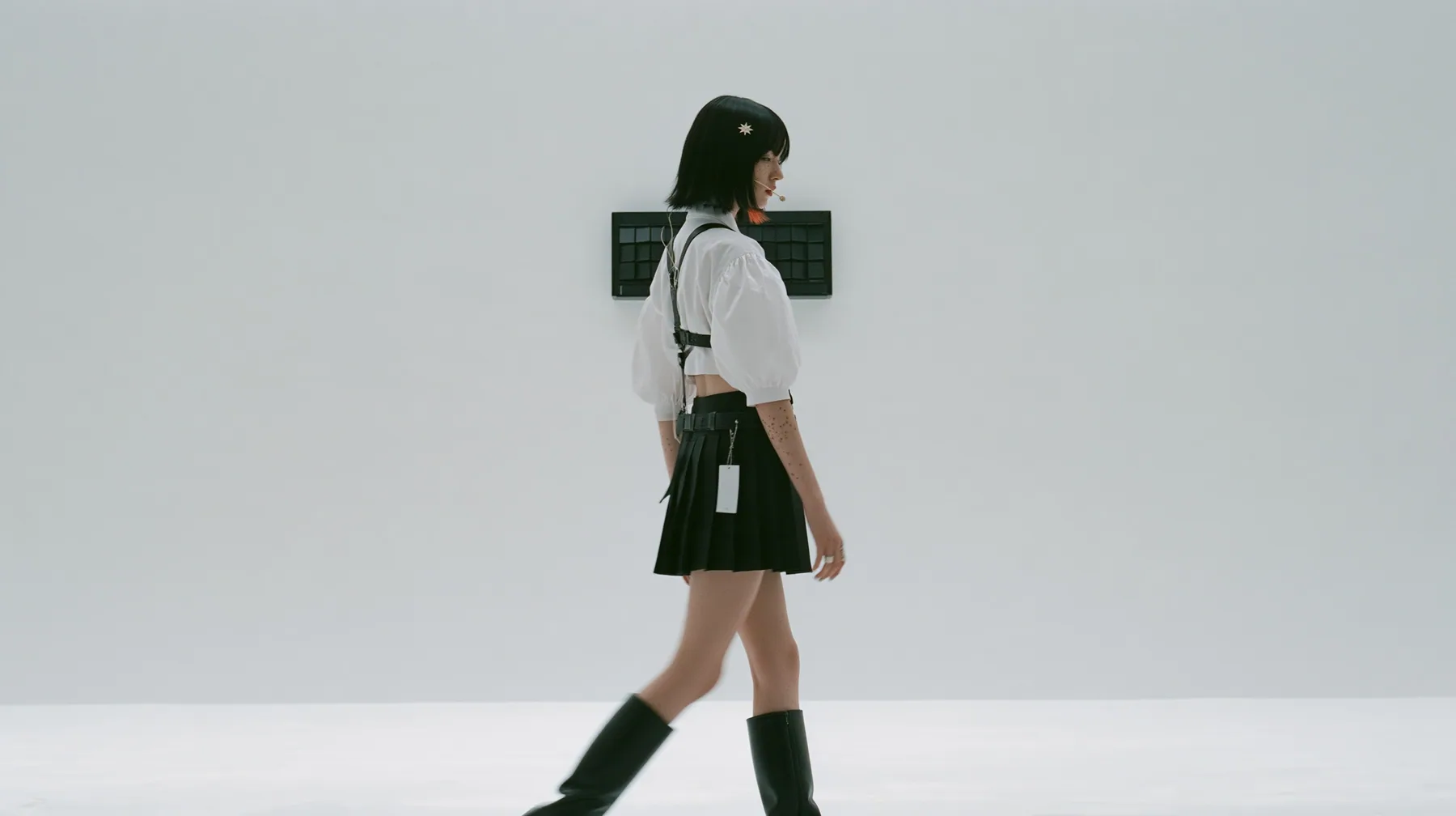 wide shot of a small black split-flap board with blank flaps alone on a seamless white paper wall, a young Caucasian American woman with pale skin and light freckles, a glossy black blunt jaw-length bob with heavy…