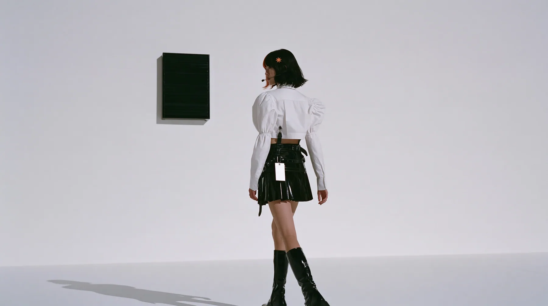 wide shot of a small black split-flap board with blank flaps alone on a seamless white paper wall, a young Caucasian American woman with pale skin and light freckles, a glossy black blunt jaw-length bob with heavy…