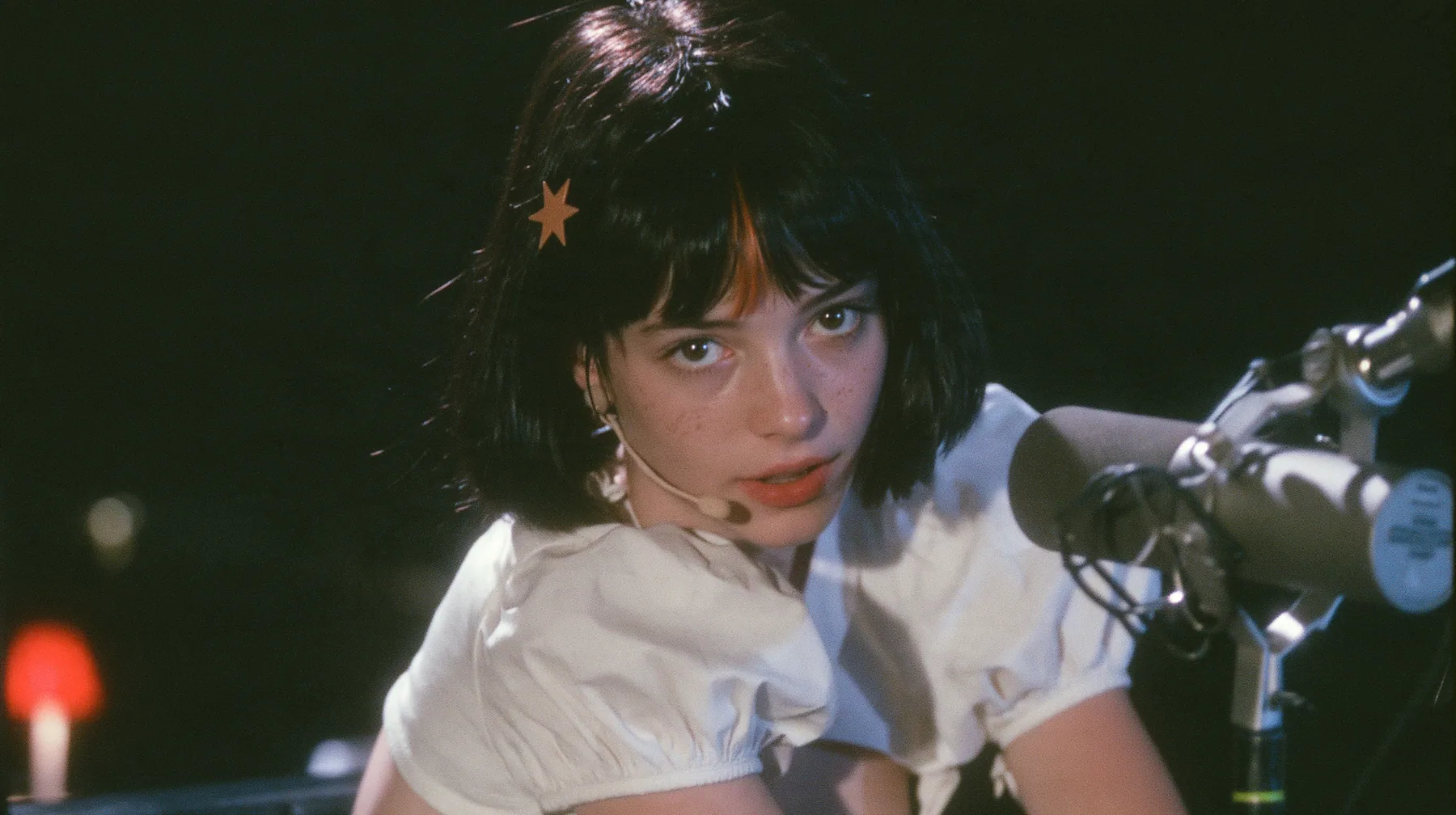 medium close-up of a young Caucasian American woman with pale skin and light freckles, a glossy black blunt jaw-length bob with heavy bangs and one clay-orange streak through the bangs, a small flat clay-orange…