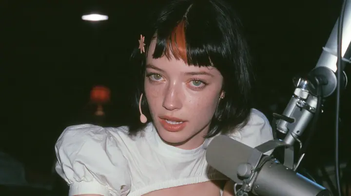 medium close-up of a young Caucasian American woman with pale skin and light freckles, a glossy black blunt jaw-length bob with heavy bangs and one clay-orange streak through the bangs, a small flat clay-orange…
