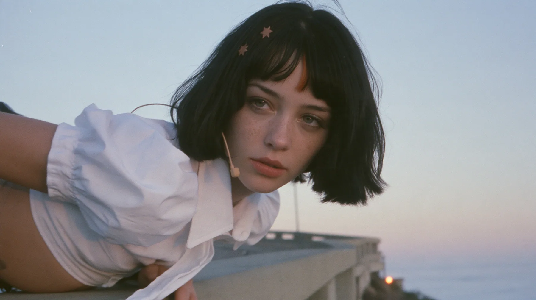 medium close-up of a young Caucasian American woman with pale skin and light freckles, a glossy black blunt jaw-length bob with heavy bangs and one clay-orange streak through the bangs, a small flat clay-orange…