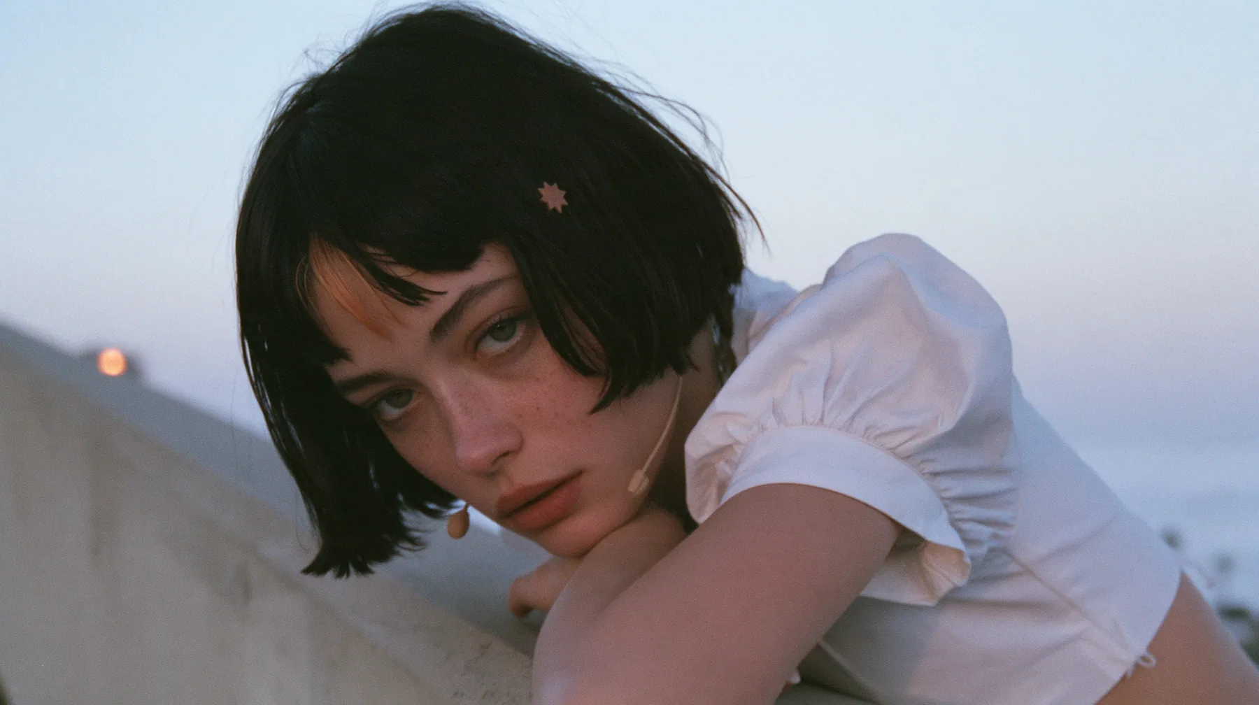 medium close-up of a young Caucasian American woman with pale skin and light freckles, a glossy black blunt jaw-length bob with heavy bangs and one clay-orange streak through the bangs, a small flat clay-orange…