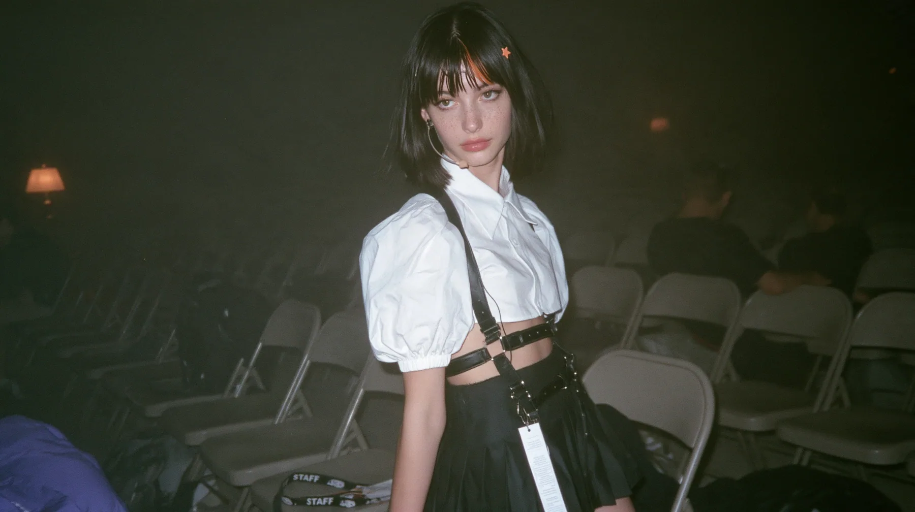 medium shot of a young Caucasian American woman with pale skin and light freckles, a glossy black blunt jaw-length bob with heavy bangs and one clay-orange streak through the bangs, a small flat clay-orange…