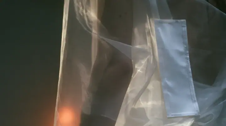 close-up of a blank white garment tag stitched to a sheer organza sleeve, the arm visible through the transparent fabric, no face, the left half empty grey, pre-dawn slate grey, soft fog, one small ember-orange lamp…