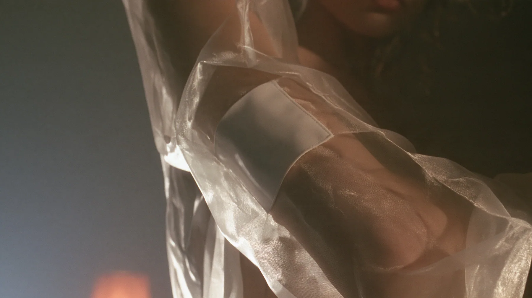 close-up of a blank white garment tag stitched to a sheer organza sleeve, the arm visible through the transparent fabric, no face, the left half empty grey, pre-dawn slate grey, soft fog, one small ember-orange lamp…