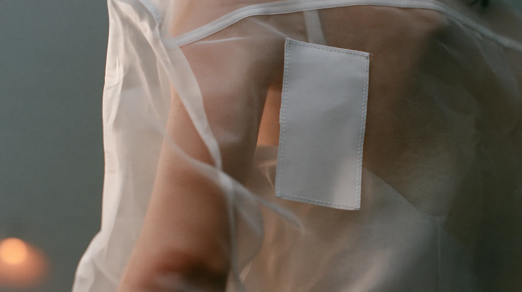 close-up of a blank white garment tag stitched to a sheer organza sleeve, the arm visible through the transparent fabric, no face, the left half empty grey, pre-dawn slate grey, soft fog, one small ember-orange lamp…