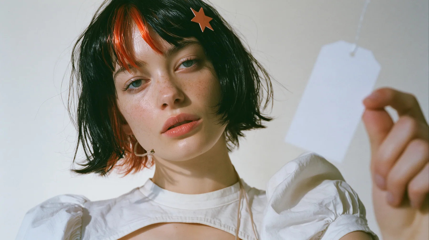 medium close-up of a young Caucasian American woman with pale skin and light freckles, a glossy black blunt jaw-length bob with heavy bangs and one clay-orange streak through the bangs, a small flat clay-orange…