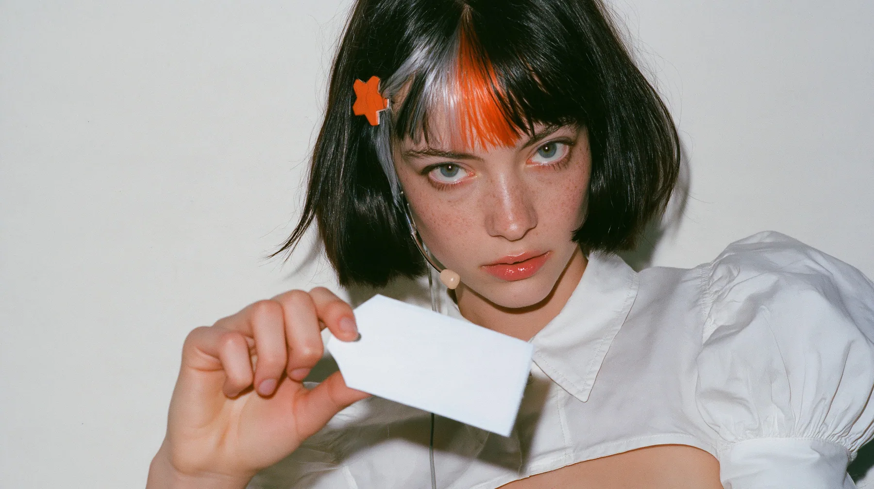 medium close-up of a young Caucasian American woman with pale skin and light freckles, a glossy black blunt jaw-length bob with heavy bangs and one clay-orange streak through the bangs, a small flat clay-orange…