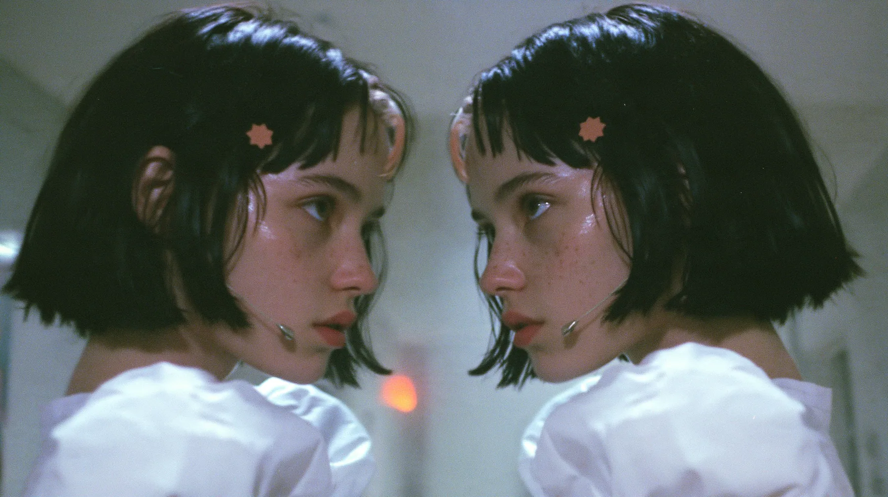 medium close-up of two identical copies of a young Caucasian American woman with pale skin and light freckles, a glossy black blunt jaw-length bob with heavy bangs and one clay-orange streak through the bangs, a…