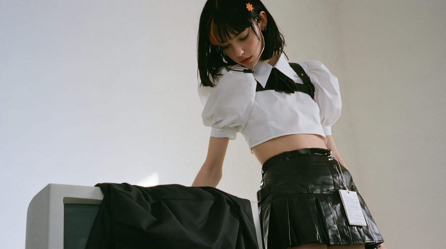 medium shot of a young Caucasian American woman with pale skin and light freckles, a glossy black blunt jaw-length bob with heavy bangs and one clay-orange streak through the bangs, a small flat clay-orange…