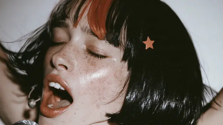 close-up of a young Caucasian American woman with pale skin and light freckles, a glossy black blunt jaw-length bob with heavy bangs and one clay-orange streak through the bangs, a small flat clay-orange…