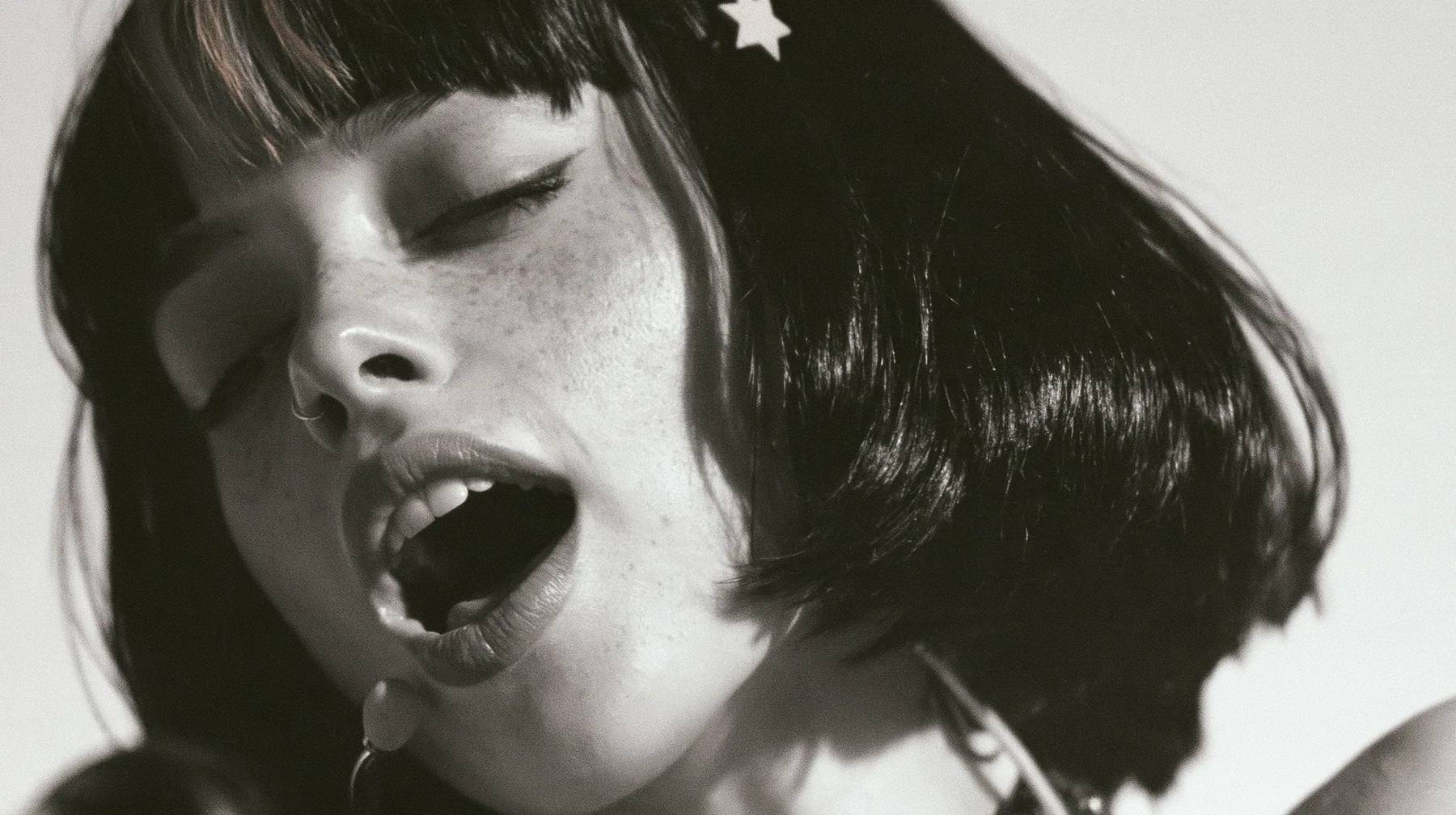 close-up of a young Caucasian American woman with pale skin and light freckles, a glossy black blunt jaw-length bob with heavy bangs and one clay-orange streak through the bangs, a small flat clay-orange…