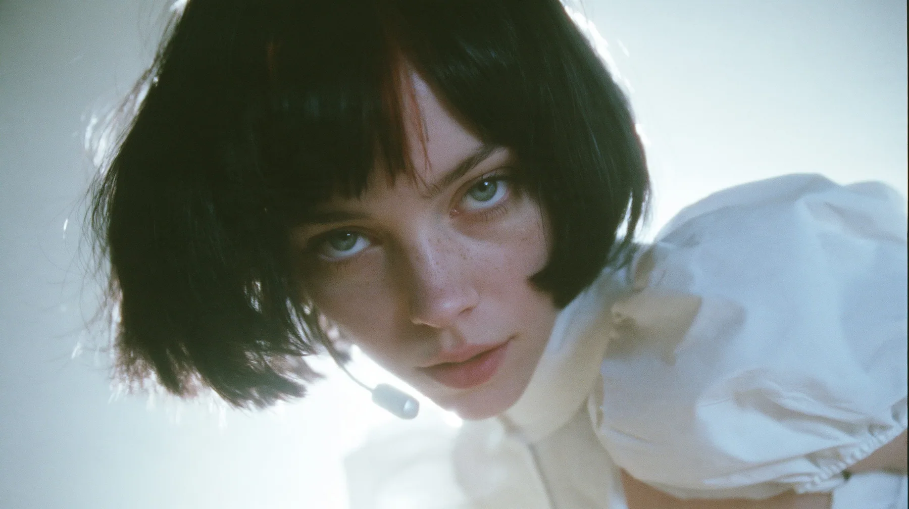 medium close-up of a young Caucasian American woman with pale skin and light freckles, a glossy black blunt jaw-length bob with heavy bangs and one clay-orange streak through the bangs, a small flat clay-orange…