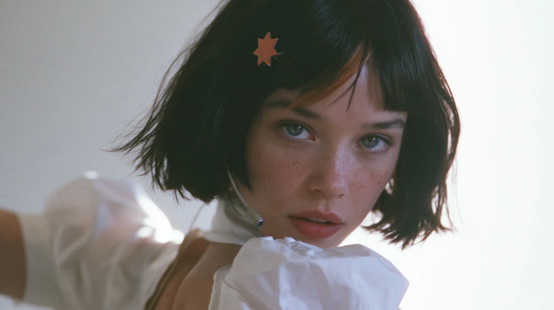 medium close-up of a young Caucasian American woman with pale skin and light freckles, a glossy black blunt jaw-length bob with heavy bangs and one clay-orange streak through the bangs, a small flat clay-orange…