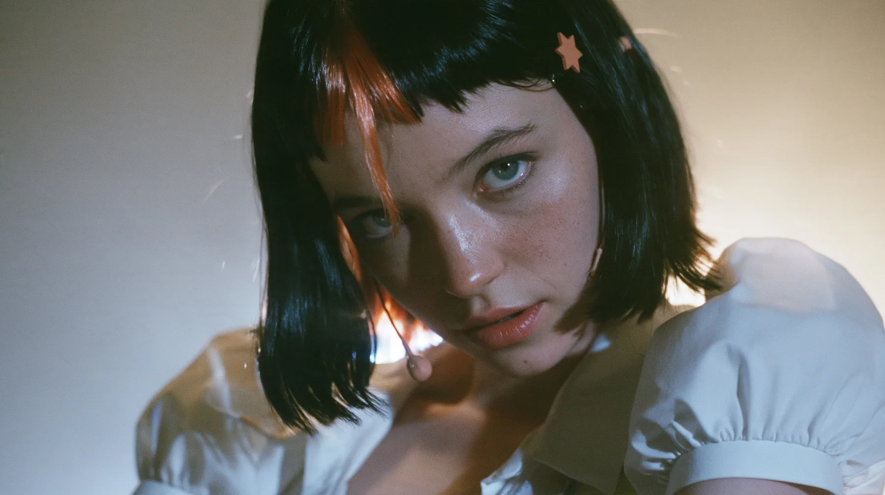 medium close-up of a young Caucasian American woman with pale skin and light freckles, a glossy black blunt jaw-length bob with heavy bangs and one clay-orange streak through the bangs, a small flat clay-orange…