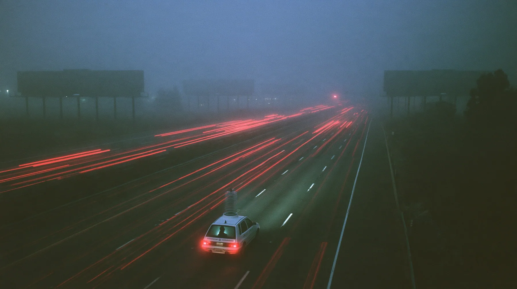 extreme wide shot from a high overpass of a fogged eight-lane freeway at night, rows of enormous blank billboards on steel poles receding into the fog, streams of red tail lights, one small white compact electric SUV…