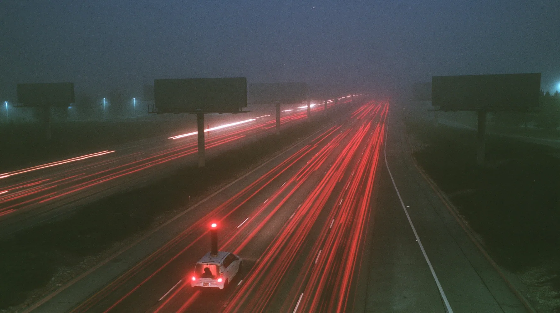 extreme wide shot from a high overpass of a fogged eight-lane freeway at night, rows of enormous blank billboards on steel poles receding into the fog, streams of red tail lights, one small white compact electric SUV…