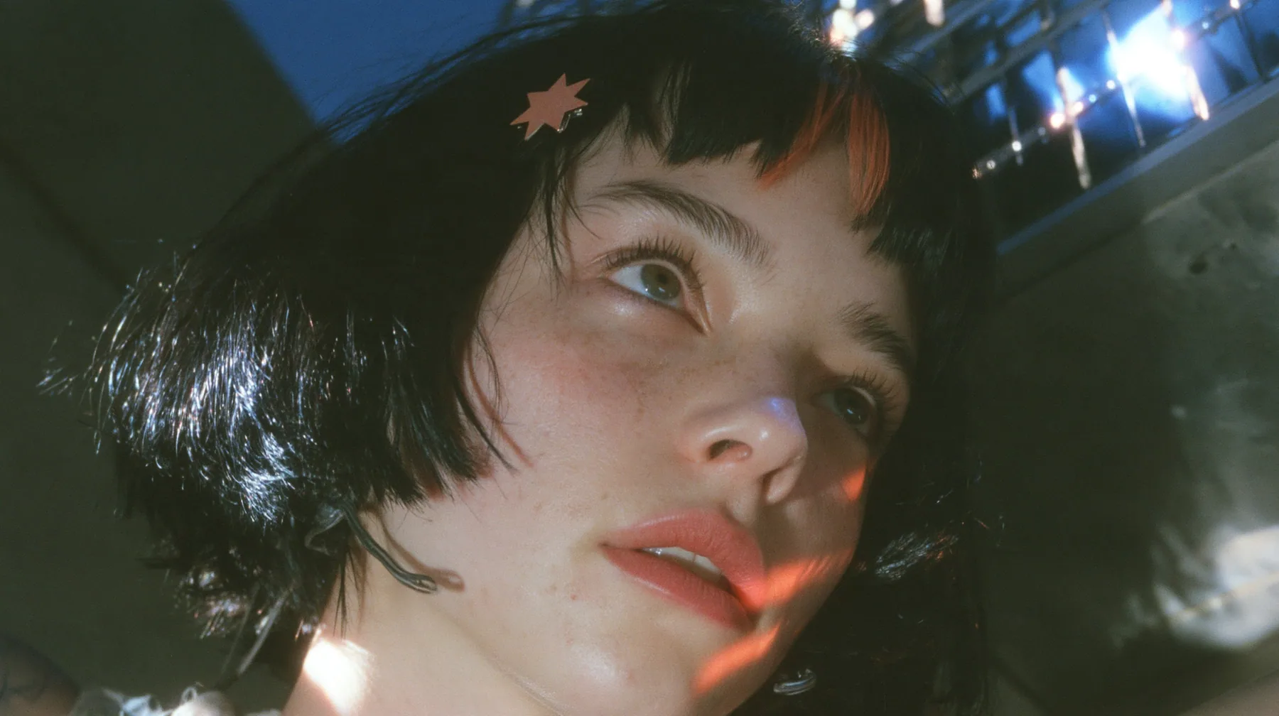 close-up from a low angle of a young Caucasian American woman with pale skin and light freckles, a glossy black blunt jaw-length bob with heavy bangs and one clay-orange streak through the bangs, a small flat…