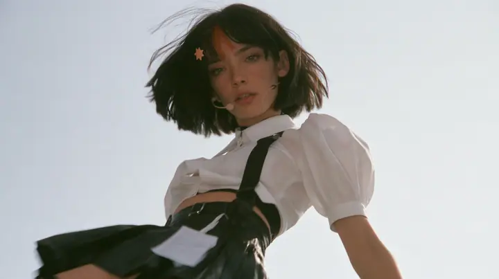 medium shot of a young Caucasian American woman with pale skin and light freckles, a glossy black blunt jaw-length bob with heavy bangs and one clay-orange streak through the bangs, a small flat clay-orange…
