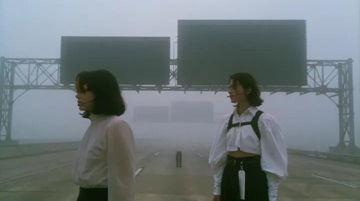 extreme wide shot of three enormous split-flap signs with blank black flaps on steel gantries spanning an empty freeway in white fog, receding one behind the other, a young Caucasian American woman with pale skin and…