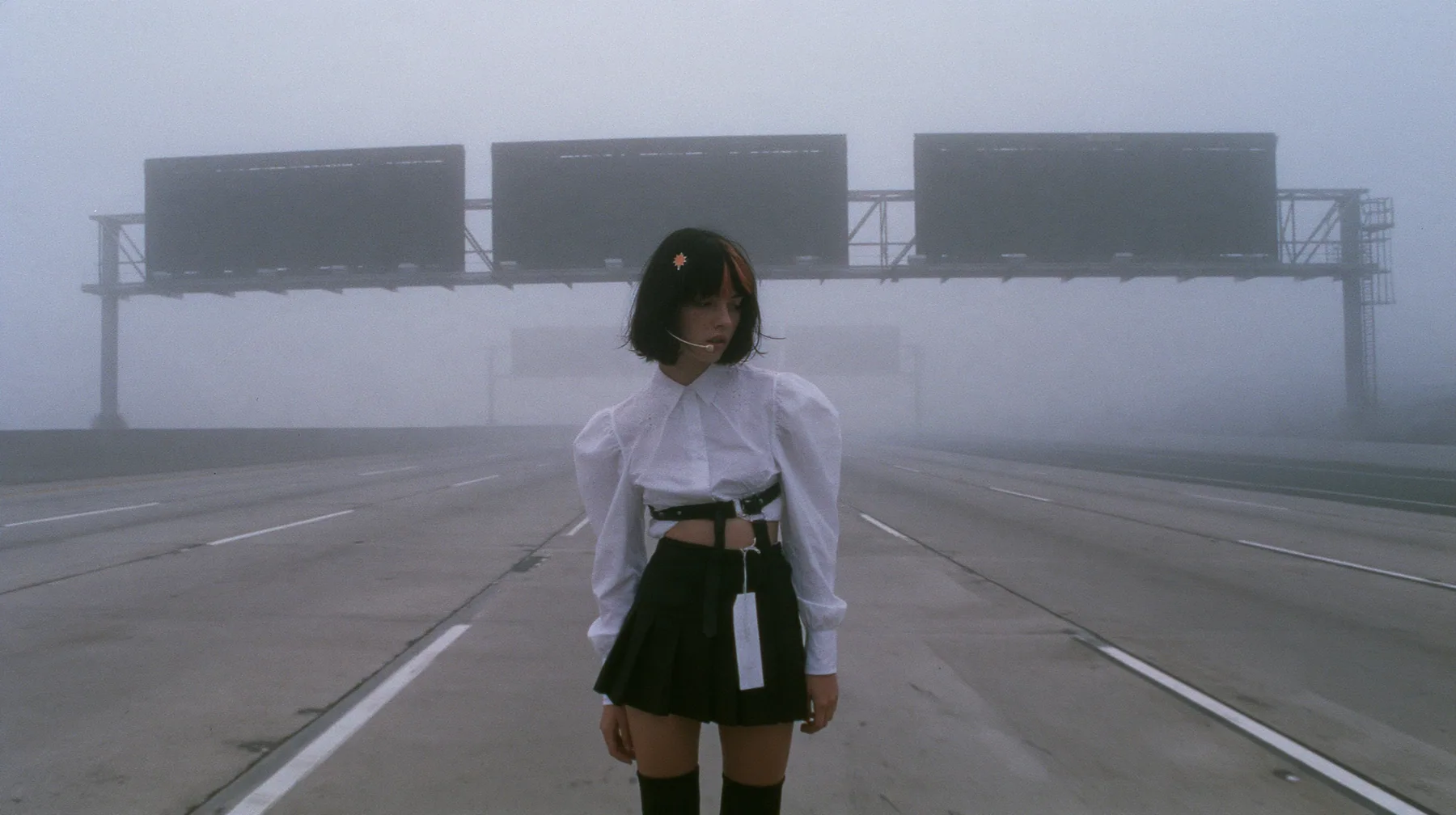 extreme wide shot of three enormous split-flap signs with blank black flaps on steel gantries spanning an empty freeway in white fog, receding one behind the other, a young Caucasian American woman with pale skin and…