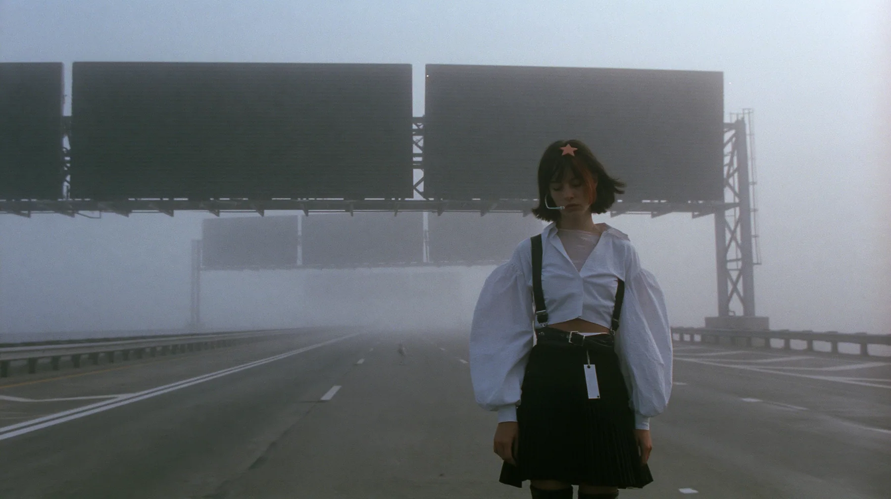 extreme wide shot of three enormous split-flap signs with blank black flaps on steel gantries spanning an empty freeway in white fog, receding one behind the other, a young Caucasian American woman with pale skin and…