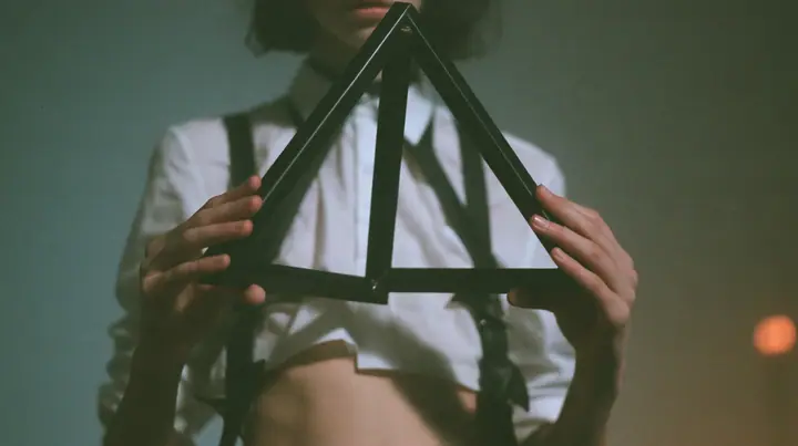 close-up of a young Caucasian American woman's hands holding an impossible triangle object made of matte black metal at chest height, a white cropped shirt and harness straps behind, no face, the right third empty…