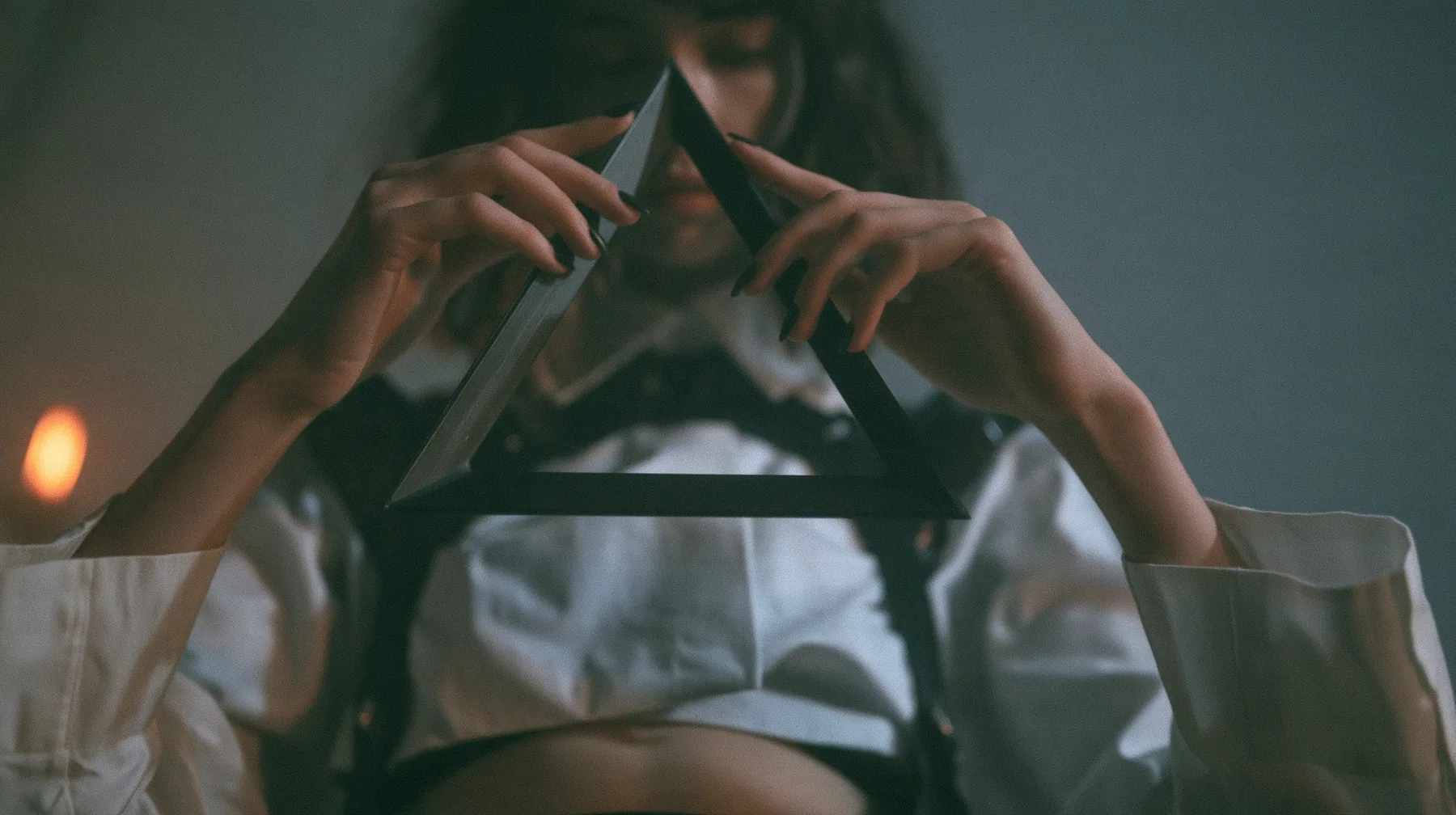 close-up of a young Caucasian American woman's hands holding an impossible triangle object made of matte black metal at chest height, a white cropped shirt and harness straps behind, no face, the right third empty…