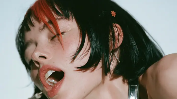 close-up in three-quarter profile of a young Caucasian American woman with pale skin and light freckles, a glossy black blunt jaw-length bob with heavy bangs and one clay-orange streak through the bangs, a small flat…