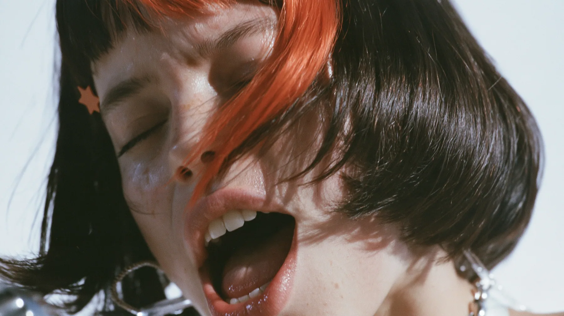 close-up in three-quarter profile of a young Caucasian American woman with pale skin and light freckles, a glossy black blunt jaw-length bob with heavy bangs and one clay-orange streak through the bangs, a small flat…