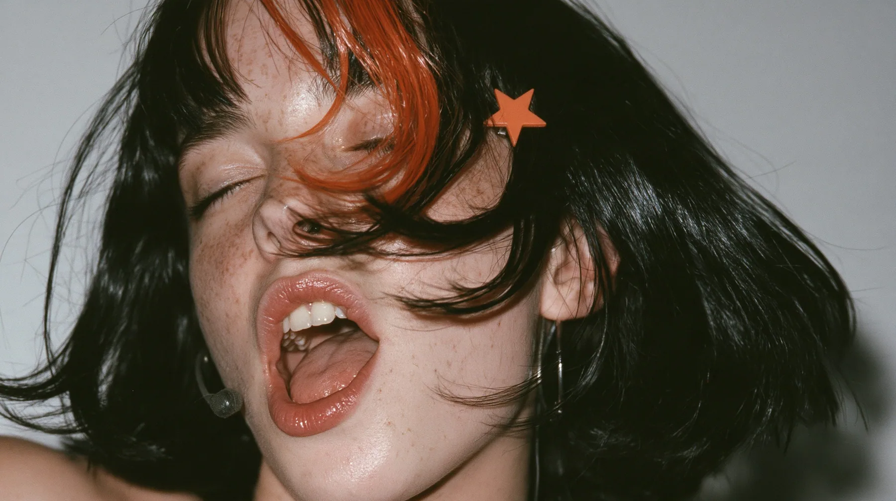 close-up in three-quarter profile of a young Caucasian American woman with pale skin and light freckles, a glossy black blunt jaw-length bob with heavy bangs and one clay-orange streak through the bangs, a small flat…