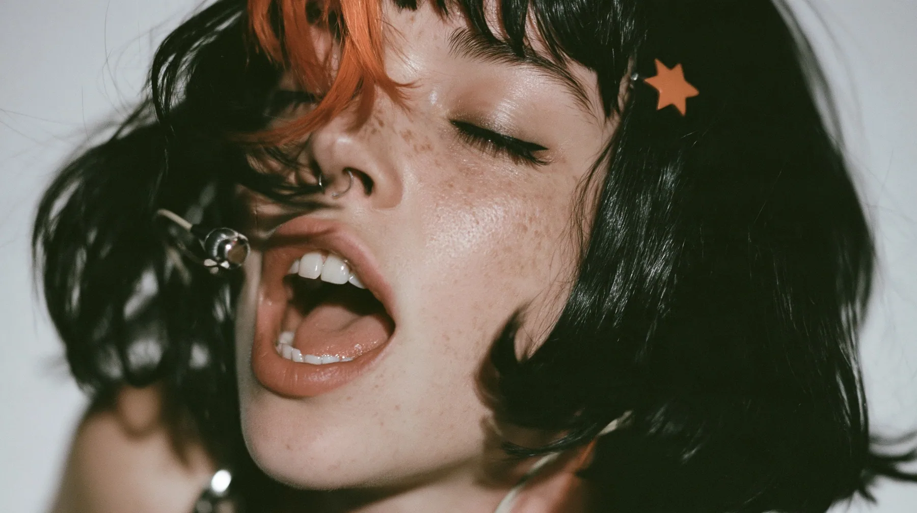 close-up in three-quarter profile of a young Caucasian American woman with pale skin and light freckles, a glossy black blunt jaw-length bob with heavy bangs and one clay-orange streak through the bangs, a small flat…