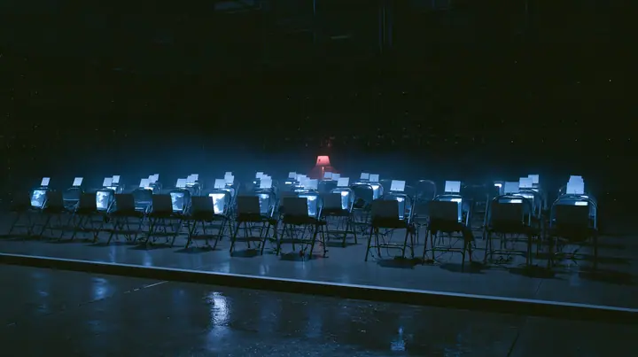 wide shot of the front row of a fashion show inside a dark server hall, every folding chair holds an open laptop glowing softly instead of a person, a small blank white card propped on each chair, the edge of the wet…