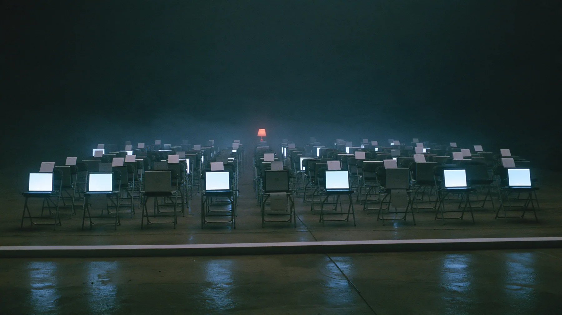 wide shot of the front row of a fashion show inside a dark server hall, every folding chair holds an open laptop glowing softly instead of a person, a small blank white card propped on each chair, the edge of the wet…