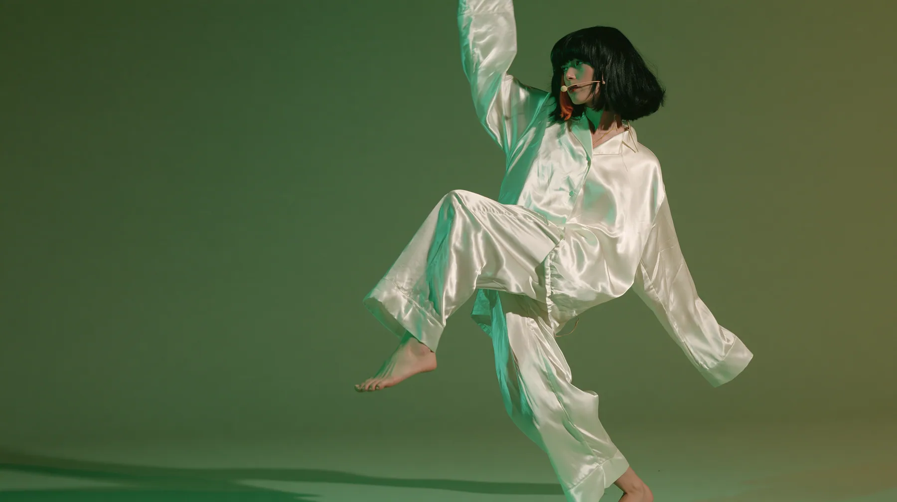 full-length studio photograph, a Caucasian American woman in her late twenties with pale skin and a glossy black blunt bob with heavy straight bangs and one clay-orange streak, a thin headset microphone, in oversized…