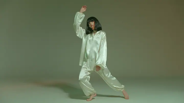 full-length studio photograph, a Caucasian American woman in her late twenties with pale skin and a glossy black blunt bob with heavy straight bangs and one clay-orange streak, a thin headset microphone, in oversized…