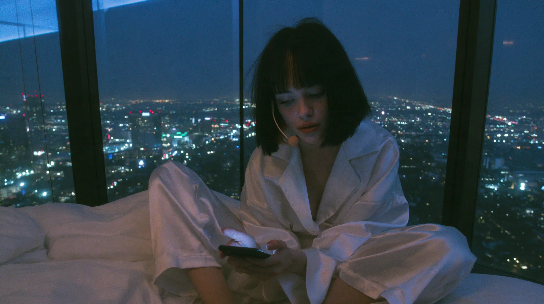 wide shot of a dark minimal high-rise bedroom at night, a floor-to-ceiling window over a glittering city, a Caucasian American woman in her late twenties with pale skin and a glossy black blunt bob with heavy…