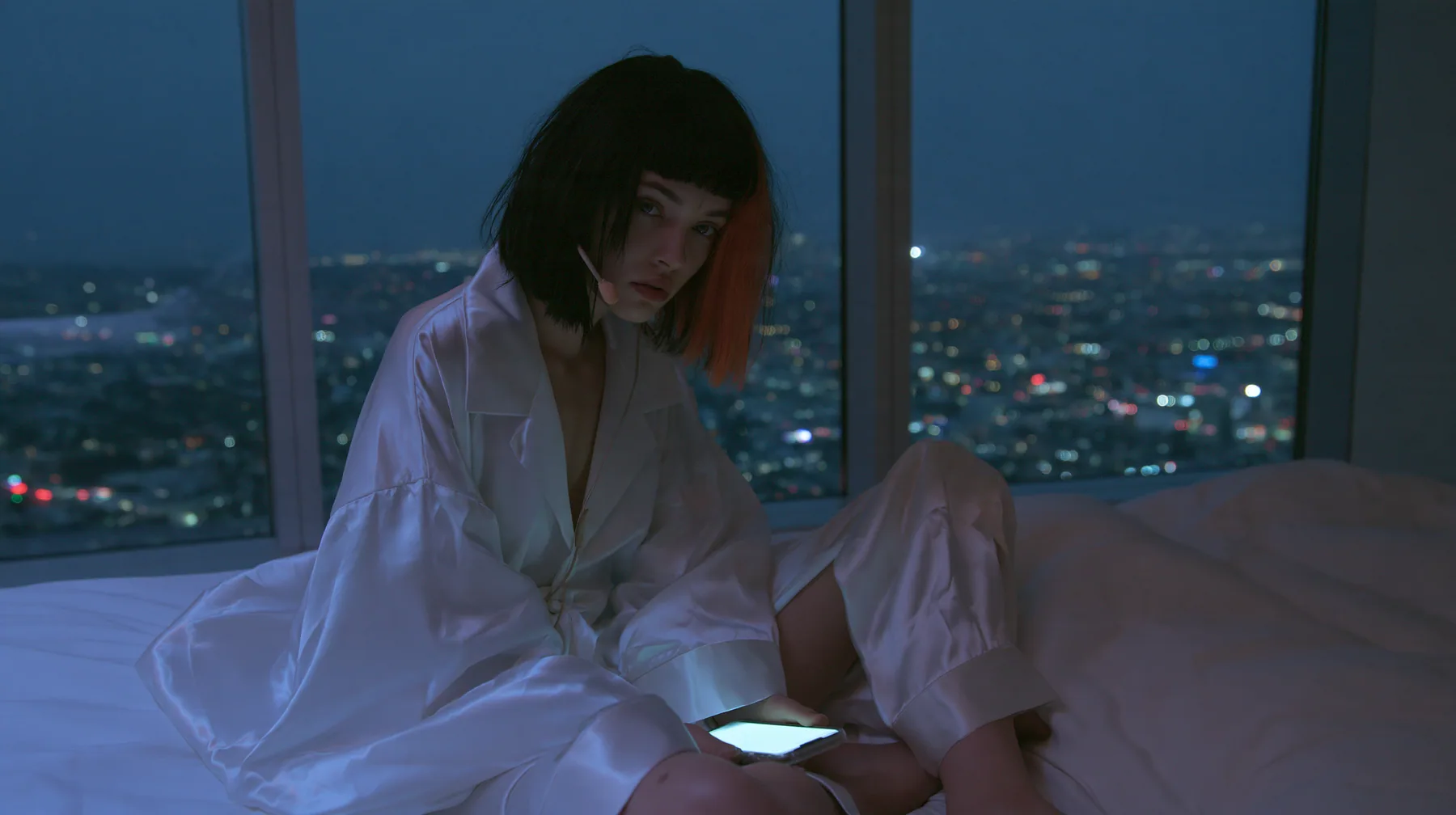 wide shot of a dark minimal high-rise bedroom at night, a floor-to-ceiling window over a glittering city, a Caucasian American woman in her late twenties with pale skin and a glossy black blunt bob with heavy…