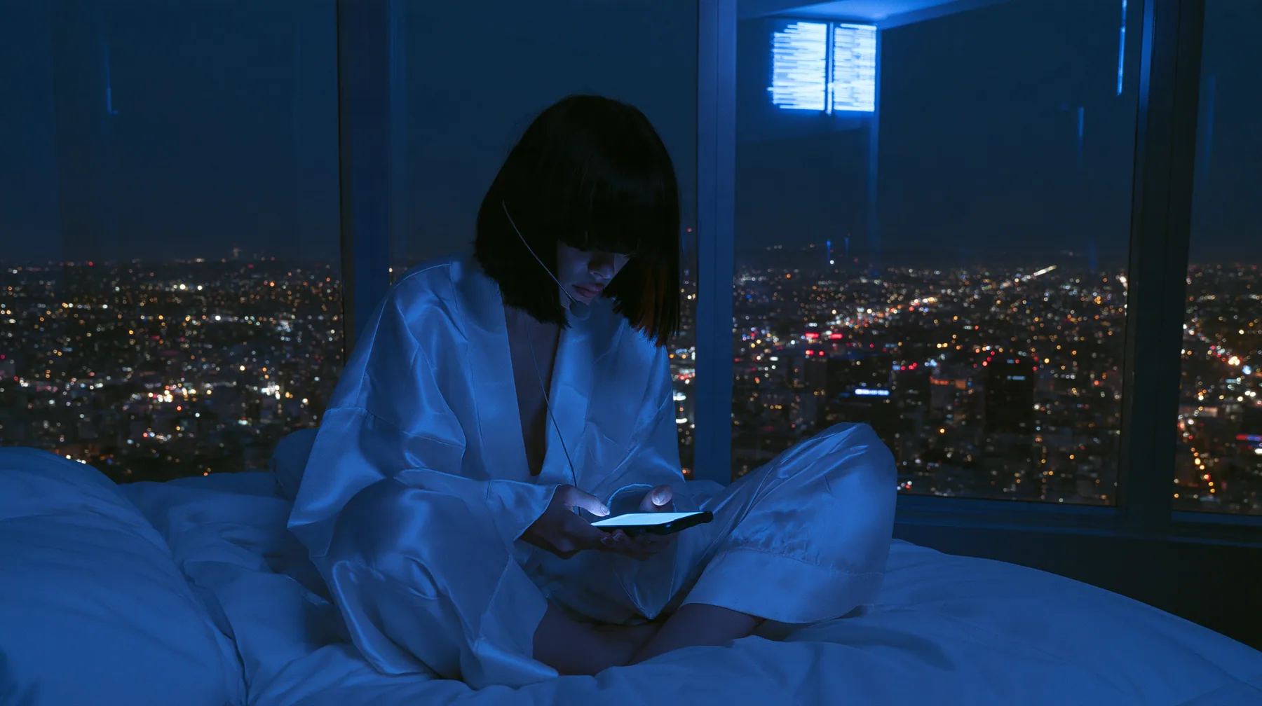 wide shot of a dark minimal high-rise bedroom at night, a floor-to-ceiling window over a glittering city, a Caucasian American woman in her late twenties with pale skin and a glossy black blunt bob with heavy…