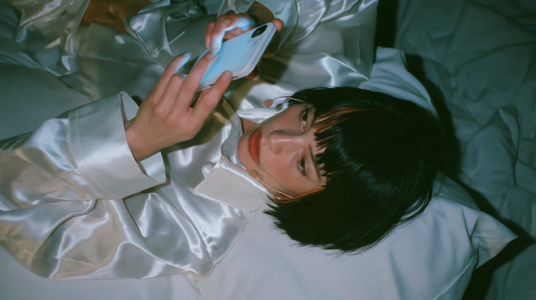 overhead shot straight down onto a white bed in the dark, a Caucasian American woman in her late twenties with pale skin and a glossy black blunt bob with heavy straight bangs and one clay-orange streak, a thin…