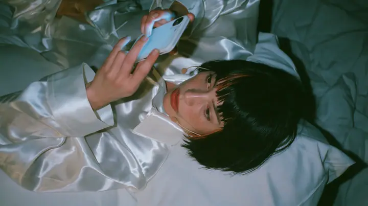 overhead shot straight down onto a white bed in the dark, a Caucasian American woman in her late twenties with pale skin and a glossy black blunt bob with heavy straight bangs and one clay-orange streak, a thin…