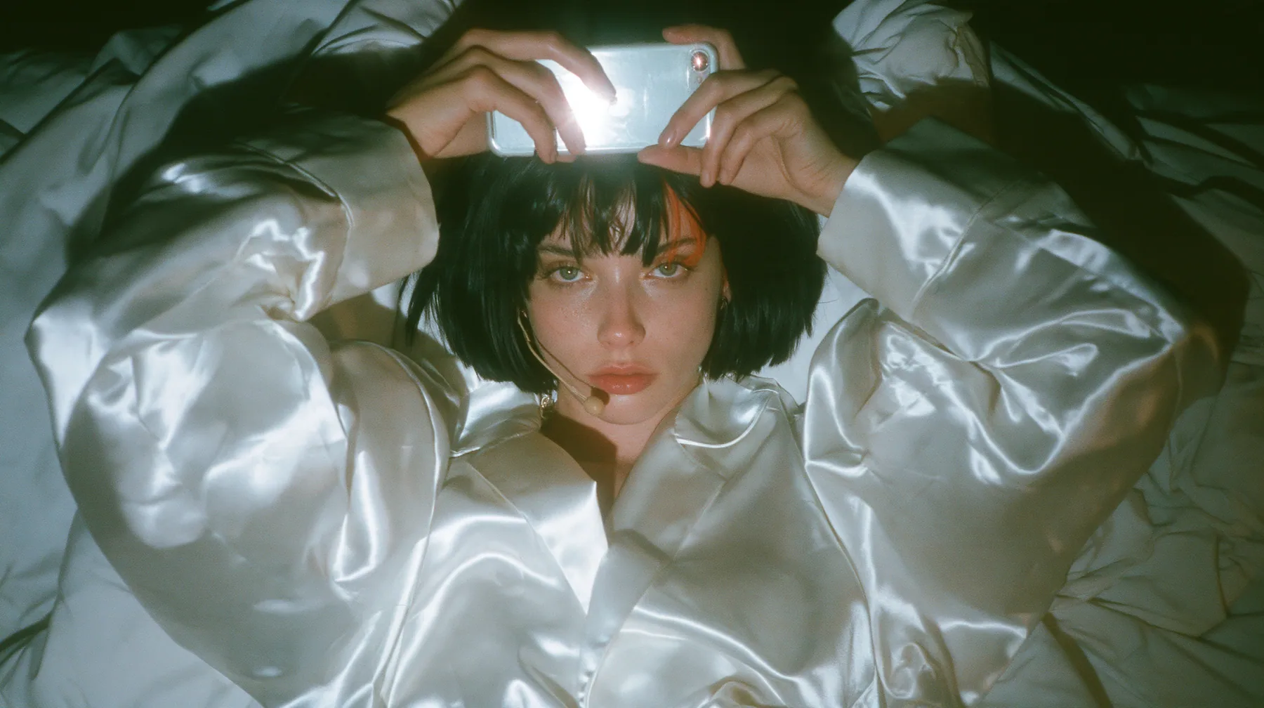 overhead shot straight down onto a white bed in the dark, a Caucasian American woman in her late twenties with pale skin and a glossy black blunt bob with heavy straight bangs and one clay-orange streak, a thin…