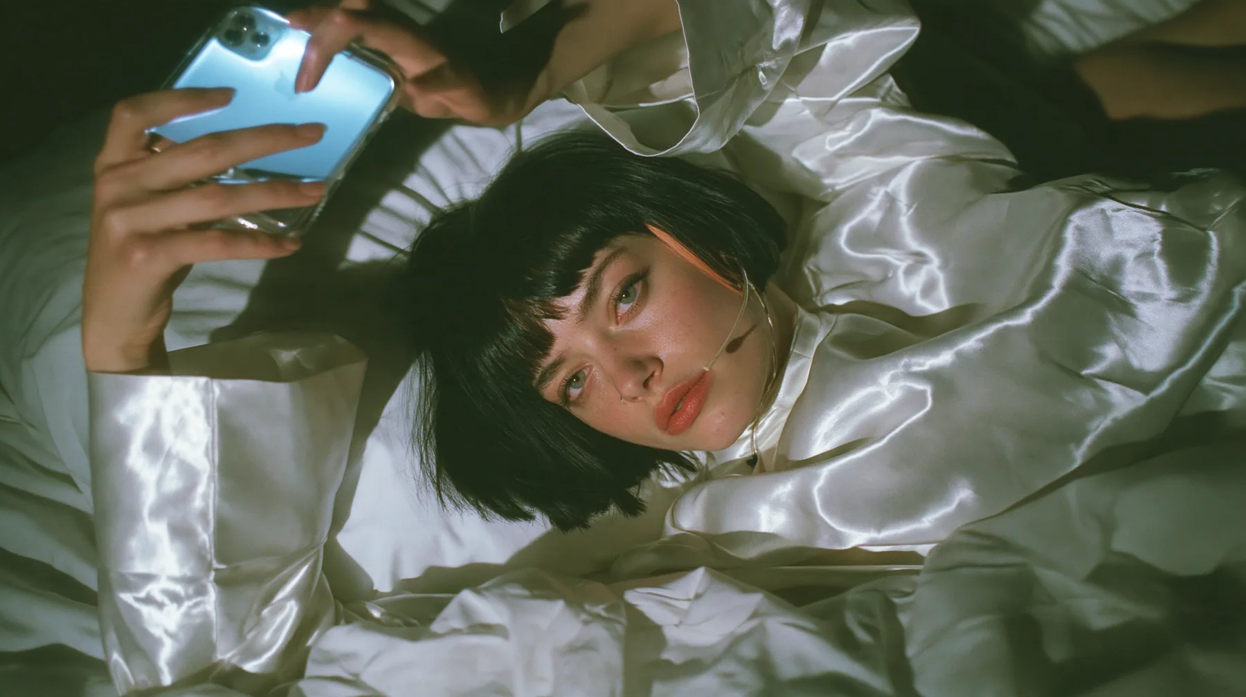 overhead shot straight down onto a white bed in the dark, a Caucasian American woman in her late twenties with pale skin and a glossy black blunt bob with heavy straight bangs and one clay-orange streak, a thin…