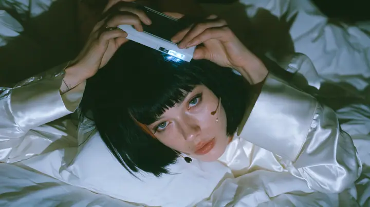 overhead shot straight down onto a white bed in the dark, a Caucasian American woman in her late twenties with pale skin and a glossy black blunt bob with heavy straight bangs and one clay-orange streak, a thin…