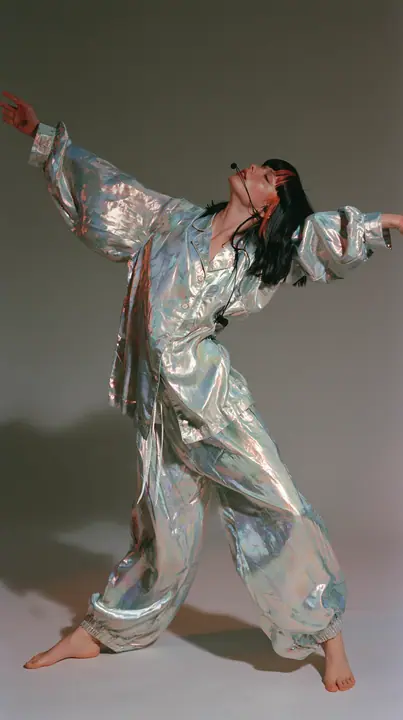 full-length shot, a Caucasian American woman in her late twenties with pale skin and a glossy black blunt bob with heavy straight bangs and one clay-orange streak, a thin headset microphone, in oversized iridescent…