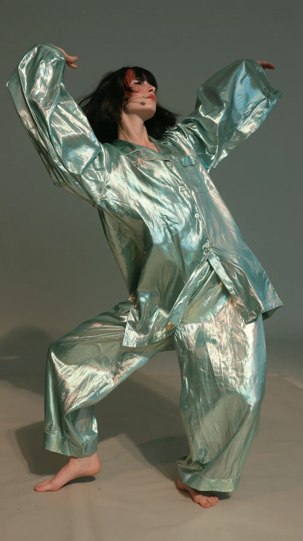 full-length shot, a Caucasian American woman in her late twenties with pale skin and a glossy black blunt bob with heavy straight bangs and one clay-orange streak, a thin headset microphone, in oversized iridescent…