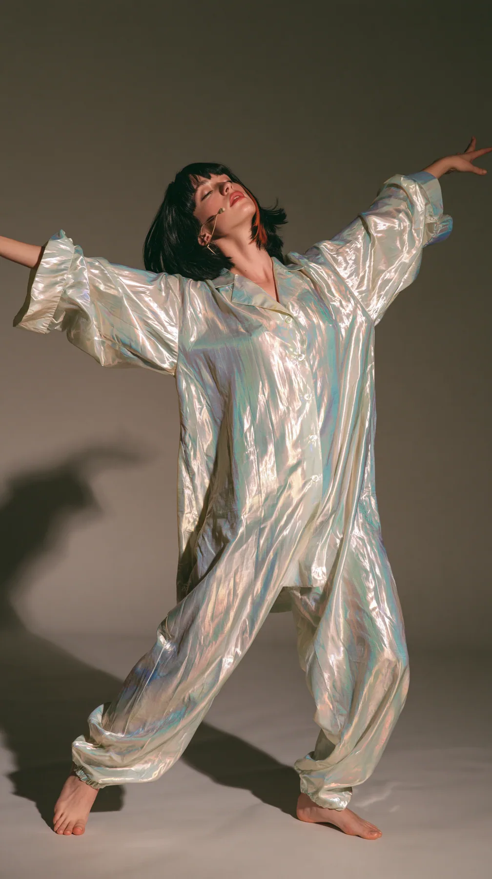 full-length shot, a Caucasian American woman in her late twenties with pale skin and a glossy black blunt bob with heavy straight bangs and one clay-orange streak, a thin headset microphone, in oversized iridescent…