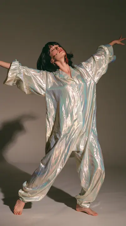 full-length shot, a Caucasian American woman in her late twenties with pale skin and a glossy black blunt bob with heavy straight bangs and one clay-orange streak, a thin headset microphone, in oversized iridescent…