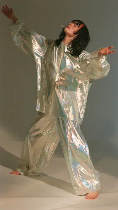 full-length shot, a Caucasian American woman in her late twenties with pale skin and a glossy black blunt bob with heavy straight bangs and one clay-orange streak, a thin headset microphone, in oversized iridescent…