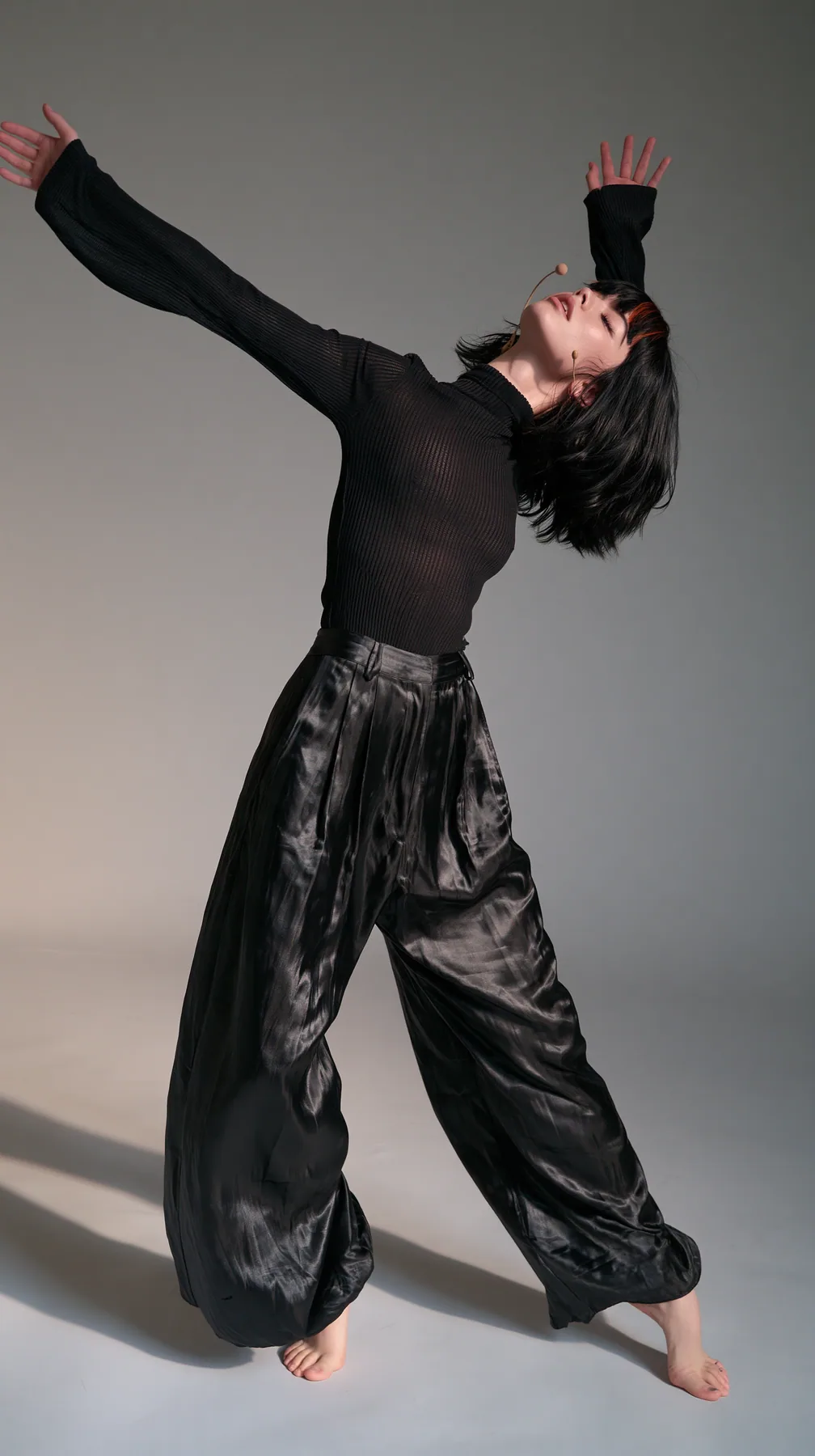 full-length shot, a Caucasian American woman in her late twenties with pale skin and a glossy black blunt bob with heavy straight bangs and one clay-orange streak, a thin headset microphone, in a black ribbed…